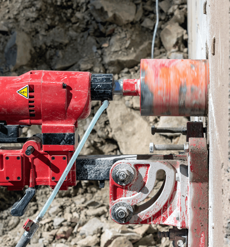 Technician operating a core drill to cut a round hole through a reinforced concrete wall.